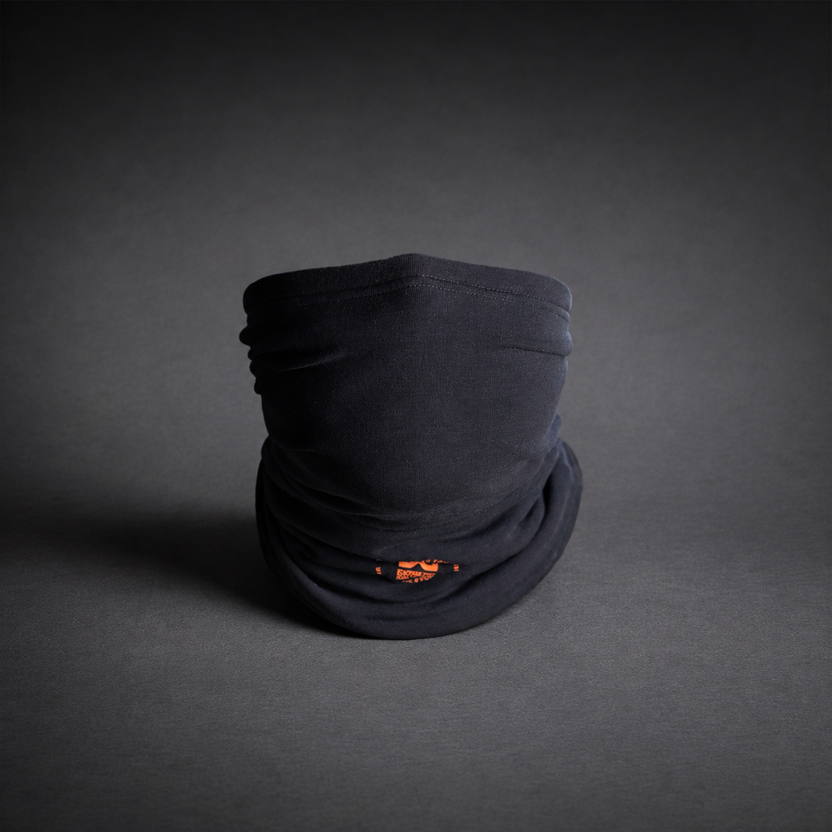 A black neck gaiter or face covering, folded and standing upright on a dark gray studio background. A small orange logo is visible near the bottom front.