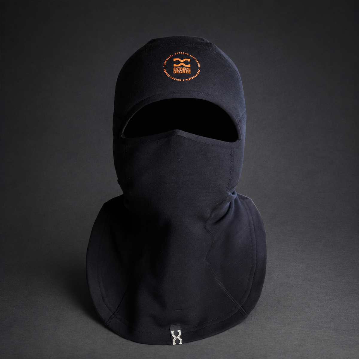 A black balaclava face mask with an integrated neck gaiter, featuring an orange embroidered logo on the forehead and a small silver metal zipper pull at the bottom, displayed on a dark gray background.